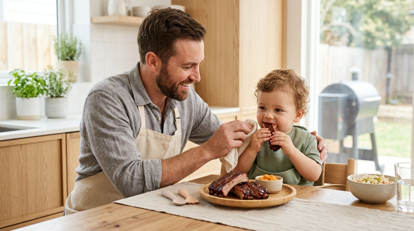 A confused dad standing next to a backyard smoker holding a baby and barbecue tongs.